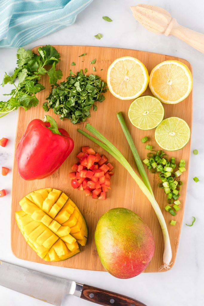 A cutting board with cilantro, lemon, red bell pepper, green onion, mango, and lime. 