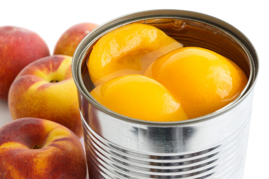 Detail of open can of peach halves in syrup on white background with whole fresh peaches.